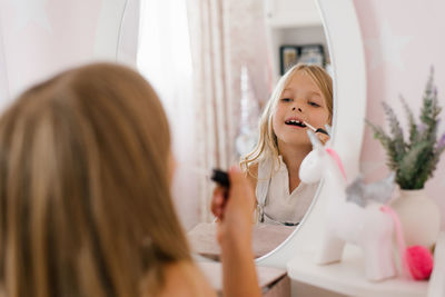 Pretty child girl paints her lips with glitter or balm in front of a mirror in her room