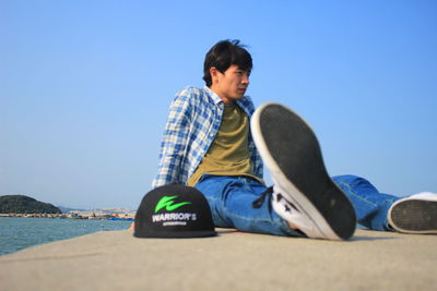 Side view of young man sitting on land against clear blue sky