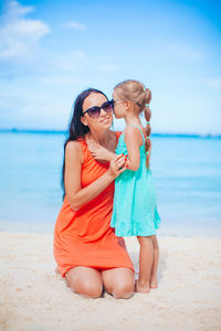 Full length of mother embracing daughter sitting on beach against sky
