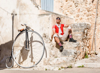 Pensive trendy male in casual clothes looking up and dreaming while sitting on exterior stairs of old building near high wheel bicycle against cloudless blue sky in sunny day