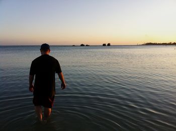 Rear view of man walking in sea against sky during sunset