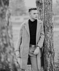 Young man looking away while standing by tree trunk