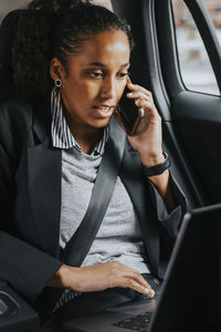 Businesswoman talking on smart phone while using laptop sitting in car