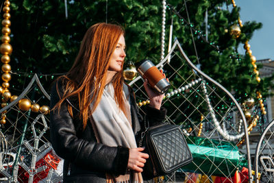 Young redhead woman drinking coffee wandering near christmas tree in the streets on winter city. 