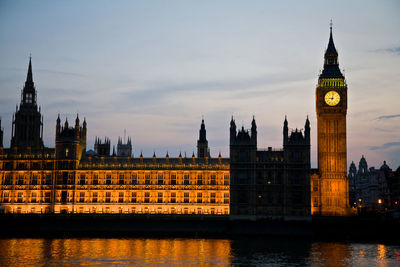 Illuminated westminster palace and big ben by thames river against sky during sunset