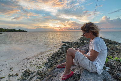 Woman sitting on rock by sea against sky during sunset