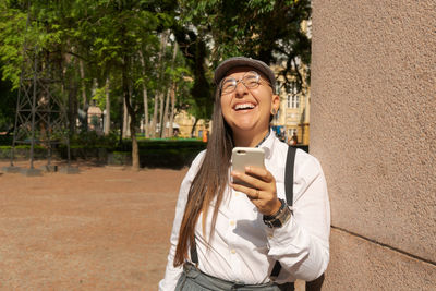 Portrait of smiling young man using phone while standing against tree