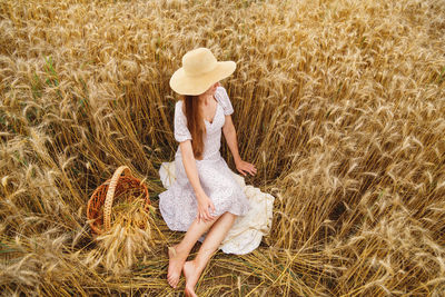 Young barefoot woman in white dress and hat sit in wheat field