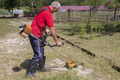 A mature man with a trimmer cuts grass and weeds with a trimmer on a paved path.