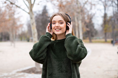 Portrait of smiling young woman listening music through headphones while standing against bare trees during winter