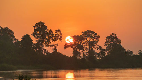 Silhouette trees by lake against orange sky