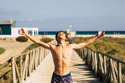Full length of shirtless man standing on railing against sky