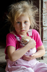  little farm girl in pink overalls hugs a small kitten  as she sits on a step outside the barn