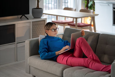 Young woman lying on comfortable sofa works on laptop and types information about business