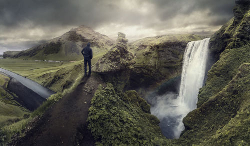 Rear view of hiker standing on rock by skogafoss against cloudy sky