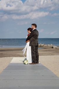 Bride and groom embracing at beach against sky