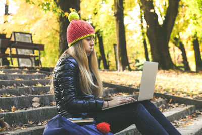 Girl with wool cap sitting in the park use laptop student teen doing homeworks with computer outdoor