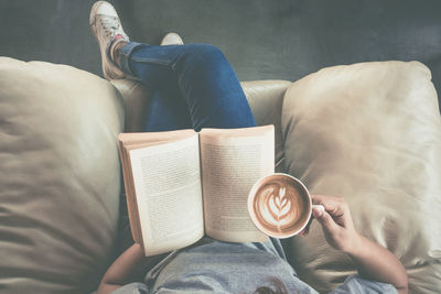 Low section of woman holding coffee cup while reading book on sofa