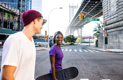 Smiling man and woman holding skateboard walking on road against bridge