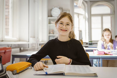 Portrait of smiling schoolgirl wearing eyeglasses sitting with book at desk in classroom