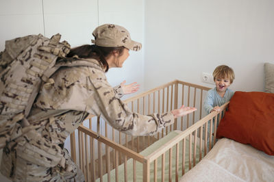 Content female soldier in uniform walking with spread arms to cute son in crib in light bedroom after mission at home