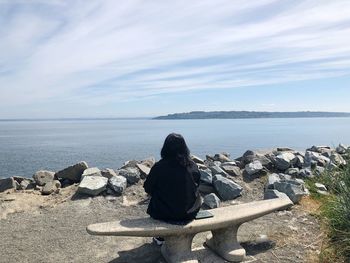 Rear view of woman sitting on rock by sea against sky