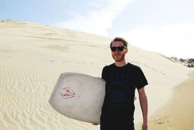Young man wearing sunglasses while standing with sandboard in dessert
