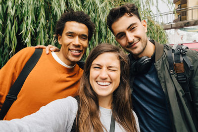 Portrait of smiling friends taking selfie while standing against trees in city