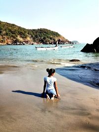 Rear view of woman kneeling on shore at beach against clear sky