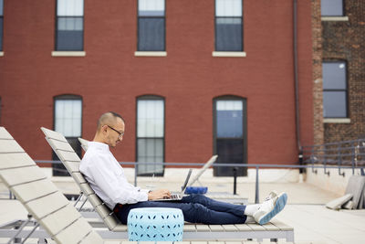 Side view of businessman using laptop while sitting on lounge chair on building terrace