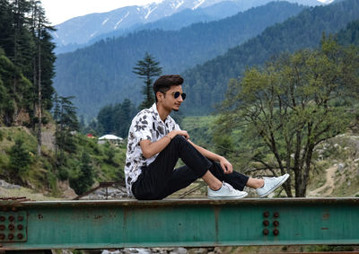 Young man sitting on railing against mountains