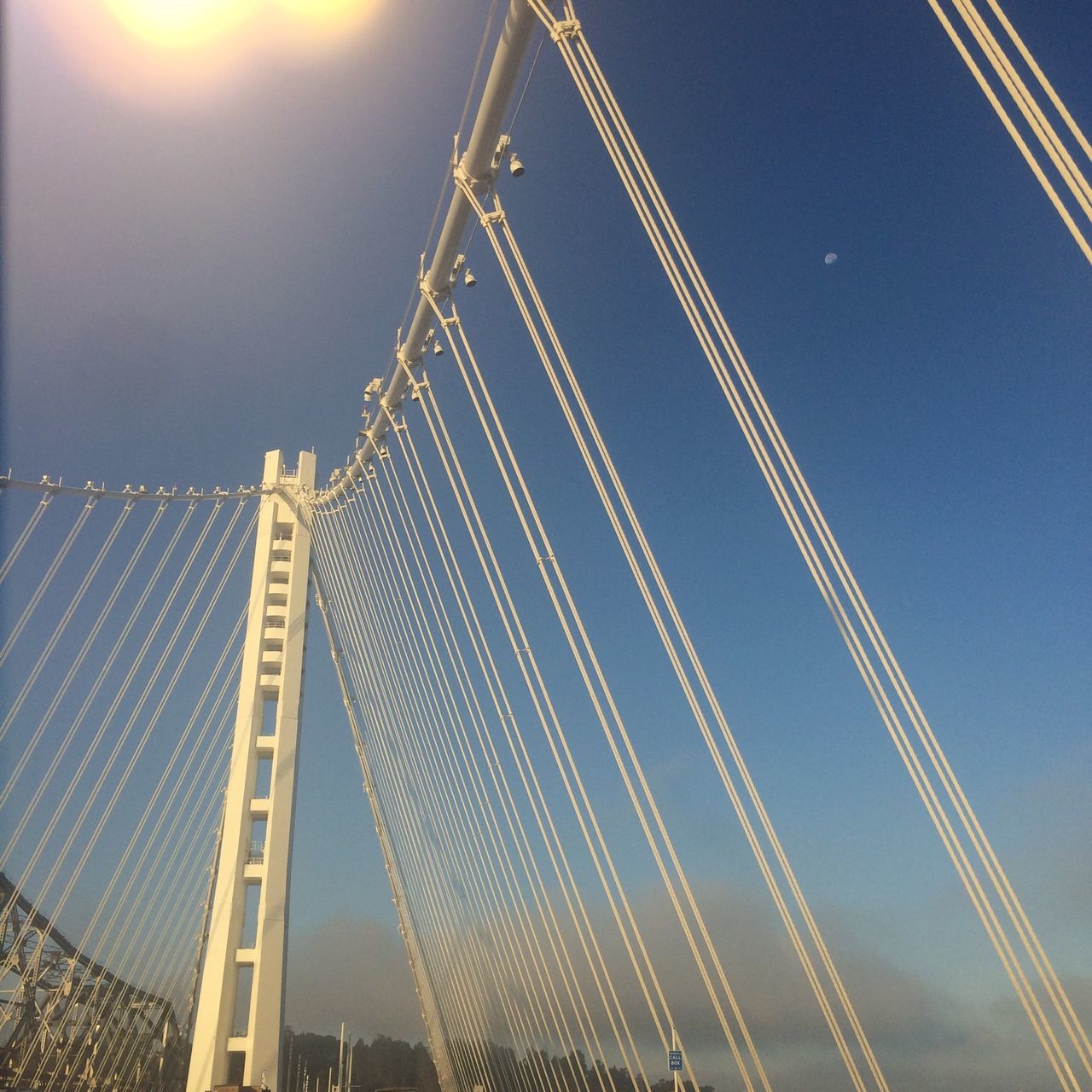 low angle view, built structure, blue, architecture, connection, sky, suspension bridge, clear sky, engineering, sunlight, tall - high, bridge - man made structure, outdoors, lens flare, transportation, cable, building exterior, cable-stayed bridge, no people, city