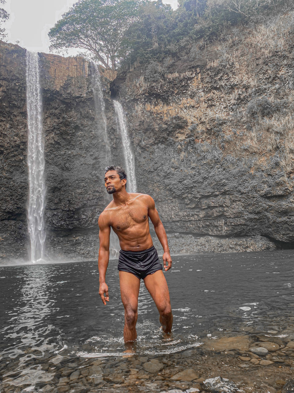 waterfall, water, one person, motion, men, nature, adult, rock, leisure activity, sports, lifestyles, scenics - nature, beauty in nature, splashing, day, holiday, vacation, land, trip, young adult, mountain, full length, outdoors, rock formation, strength, adventure, wet, muscular build, sea, travel, front view, standing, vitality, clothing, travel destinations, environment, shorts, enjoyment, activity
