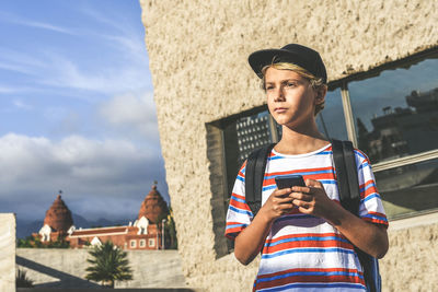 Boy holding phone while standing outside building on sunny day