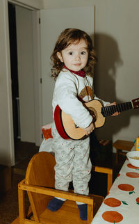 A little girl plays the guitar while standing on a chair at the table.