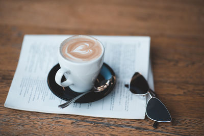 Tasty fresh cup of coffee staying on open newspaper with stylish sun glasses on wooden table in cafe