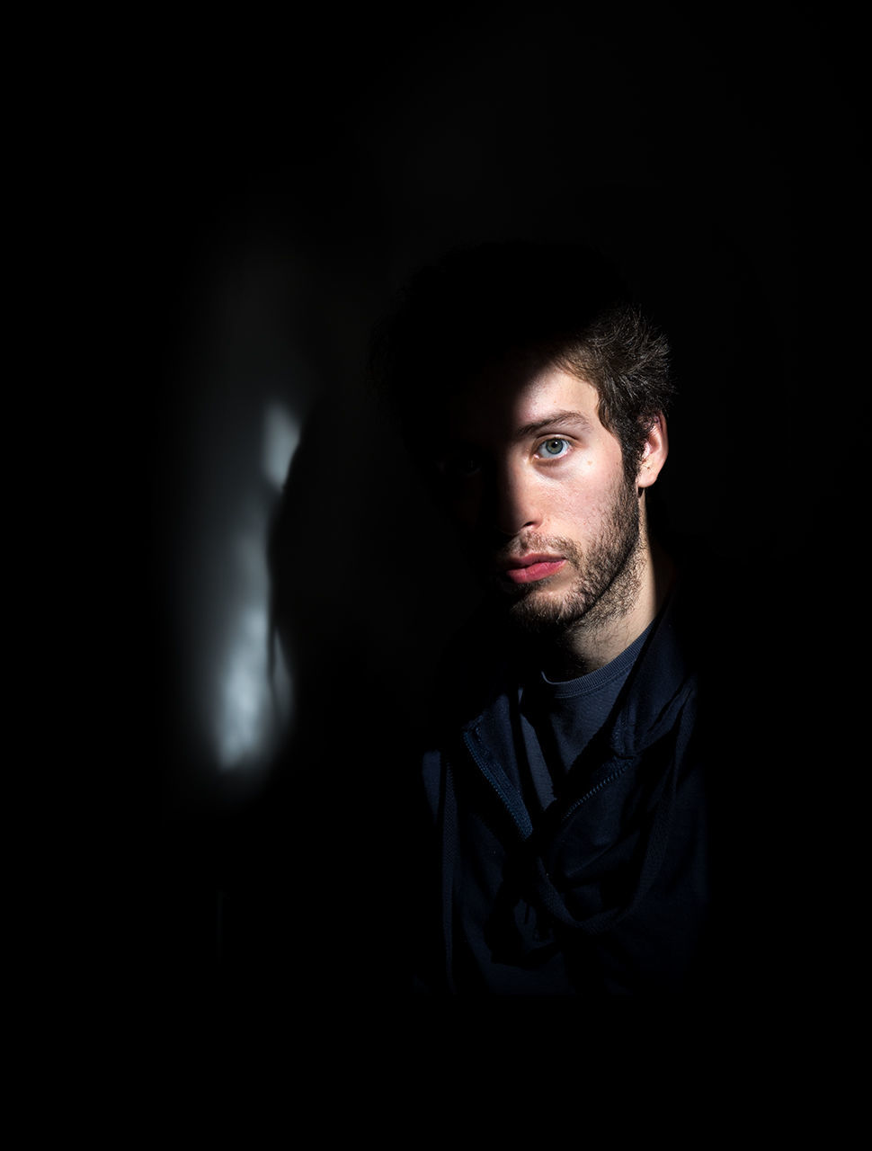 one person, portrait, beard, indoors, front view, dark, looking at camera, headshot, young adult, facial hair, young men, real people, lifestyles, men, black background, mid adult, adult, looking, contemplation, human face