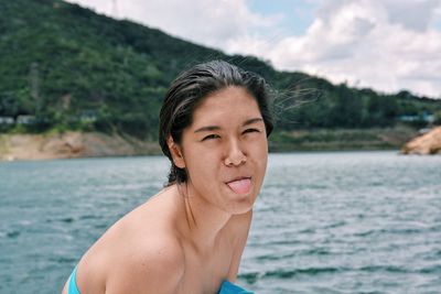 Portrait of shirtless teenage girl sticking out tongue in sea against sky