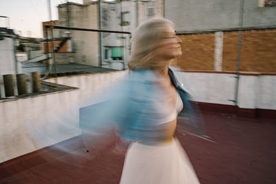 Blurred female in street style outfit moving and dancing on roof of old building