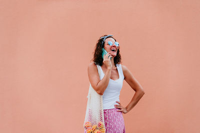 Woman with fruits talking over phone while standing on footpath against wall