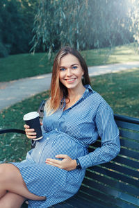 Portrait of smiling pregnant young woman sitting on bench in park