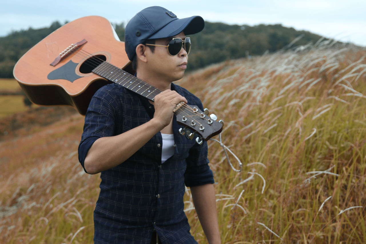guitar, music, musical instrument, three quarter length, casual clothing, plucking an instrument, hat, guitarist, standing, field, young adult, musician, one person, playing, people, grass, adult, outdoors, performance, men, artist, adults only, sky, day