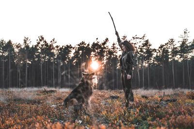 Woman holding stick with dog on field against sky