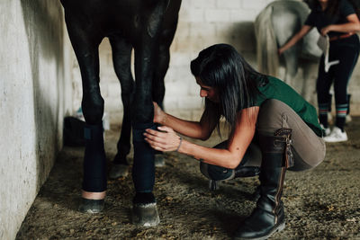 Woman examining horse's leg at stable while female friend seen in background