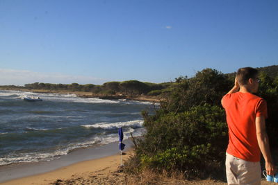 Rear view of young man standing at beach against clear sky