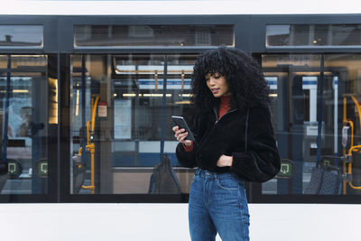 Fashionable young woman using smart phone in front of tram