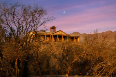 Bare trees and buildings against sky at sunset