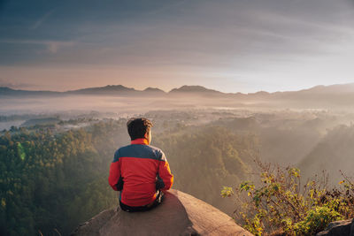 Rear view of man sitting on mountain against sky during sunset