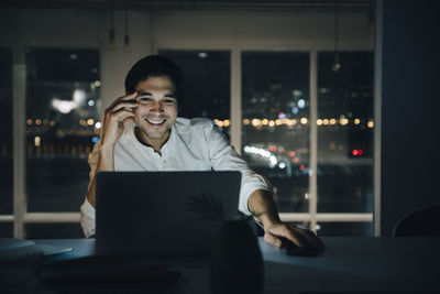 Smiling male professional working late while looking at laptop in dark coworking space