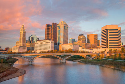 Bridge over river by buildings against sky during sunset